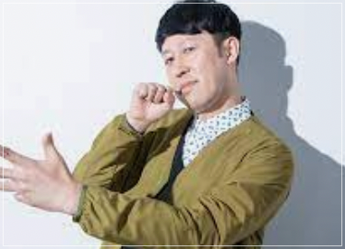 カーキの服を着てカメラ目線で歌を歌うようなポーズを取る小籔千豊さん